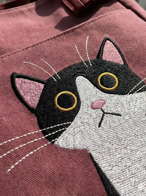 A maroon canvas tote bag featuring a large, embroidered black and white tuxedo cat with wide yellow eyes on the front pocket.