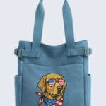 A light blue canvas tote bag featuring an embroidered Golden Retriever wearing sunglasses with an American flag lens pattern and a matching patriotic bandana