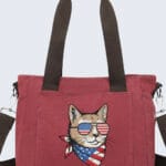 A burgundy canvas tote bag featuring an embroidered illustration of a cool cat wearing American flag sunglasses and a matching patriotic bandana. The bag has dark brown handles and a detachable shoulder strap against a clean, light background.