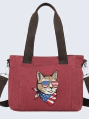 A burgundy canvas tote bag featuring an embroidered illustration of a cool cat wearing American flag sunglasses and a matching patriotic bandana. The bag has dark brown handles and a detachable shoulder strap against a clean, light background.