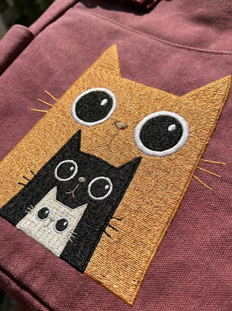 Wide-Eyed Wonder embroidery art featuring a triple-decker feline family stack in vibrant ginger, black, and white threads on a textured canvas bag.