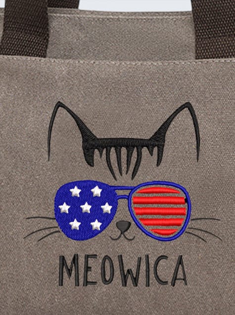 Embroidered design on a gray canvas bag featuring a cat's ears, whiskers, and sunglasses. The sunglasses have the American flag pattern with white stars on a blue lens and red stripes on the other, above the text 'MEOWICA'.