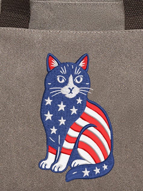 The Purr-triot: A whimsical embroidery design of a sitting cat filled with the American flag's stars and stripes, featured on a grey canvas tote bag.
