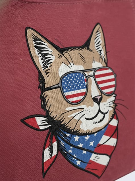 Extreme close-up of a high-quality embroidery design on a burgundy canvas tote bag featuring a cool cat wearing American flag sunglasses and a patriotic bandana.